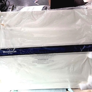 May include: A white Staedtler drafting board encased in clear plastic packaging with white foam corner protectors. The board features a dark blue horizontal drafting head with the Staedtler logo and model number printed in white on the surface.