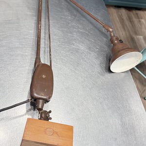May include: A brown articulated desk lamp with a wooden base. The lamp has a long, adjustable arm and a small, round lampshade with a white interior. The lamp is designed to be positioned to direct light.