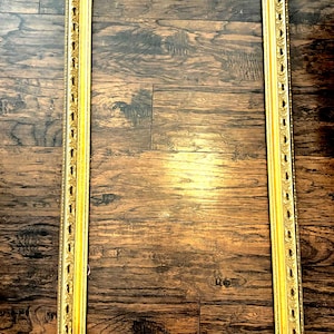 Könnte beinhalten: Ein rechteckiger, verzierter goldener Bilderrahmen mit detaillierten Schnitzereien. Der Rahmen ist leer und liegt auf einer dunklen Holzmaserung. Das Design des Rahmens weist komplizierte Muster an den Rändern und Ecken auf.