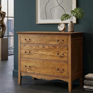 May include: A wooden chest of drawers featuring three horizontal drawers with brass-toned metal handles and central keyholes. The piece has a natural wood grain finish, a rectangular top surface, and four short legs with a scalloped apron detail.