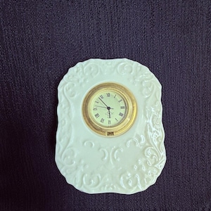 May include: A white ceramic clock with a gold-colored clock face and Roman numerals. The clock is set in a decorative, ornate frame with a floral design. The clock is approximately 4 inches wide.