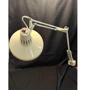 Puede incluir: Lámpara de escritorio ajustable de color crema con una pantalla redonda y un brazo giratorio. La lámpara tiene un interruptor de encendido/apagado rojo y blanco y ranuras verticales en la pantalla. La lámpara es de metal y tiene una base rectangular.