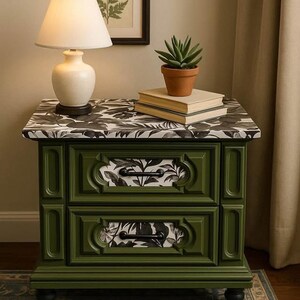 May include: A green nightstand with two drawers, black hardware, and a black and white floral pattern on the drawer fronts and tabletop. A white lamp, books, and a potted plant sit on top. A framed botanical print hangs on the wall.