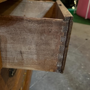 May include: A wooden dresser drawer pulled partially open, revealing the natural brown wood grain and visible dovetail joinery on the side panel. The drawer features a dark metal handle on the front face, with additional drawers visible below.