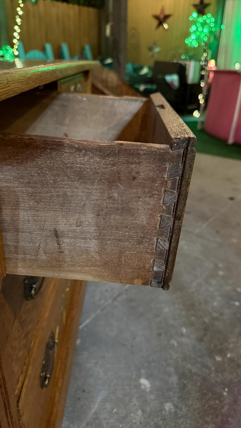 May include: A wooden dresser drawer pulled partially open, revealing the natural brown wood grain and visible dovetail joinery on the side panel. The drawer features a dark metal handle on the front face, with additional drawers visible below.