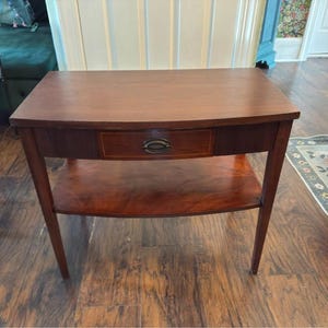 May include: A rectangular wooden side table with a smooth brown finish, featuring a single central drawer with an oval metal pull handle and a lower open shelf. The table stands on four straight legs and has a flat top surface with visible wood grain.