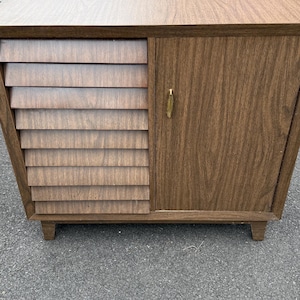 May include: A brown wooden cabinet featuring a flat top surface, a single door with a small gold-toned handle, and a side section composed of horizontal slats. The piece is supported by four short, tapered legs and has a wood grain finish.