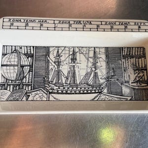 May include: A rectangular white ceramic tray featuring a black and white illustration of a sailing ship, a globe, and bookshelves. The top border displays a grid with the text ZONA TEMP. MER., ZONA TORRIDA, and ZONA TEMP. SETT. printed clearly.