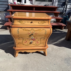 May include: A light brown, antique-style chest of drawers with a curved front. The chest features floral painted details in red, pink, and green. It has a single drawer at the top and two drawers below, with decorative hardware and a varnished finish.