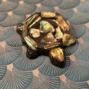 Puede incluir: Un cenicero decorativo con forma de tortuga. El cenicero es negro con detalles iridiscentes de concha de abulón alrededor del borde y dentro del cuenco. La cabeza y las patas de la tortuga también son negras con tallas detalladas.