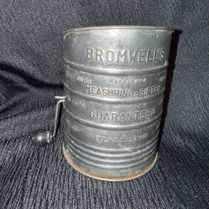 May include: A vintage metal Bromwell's measuring sifter with a cylindrical body and a side-mounted hand crank featuring a black knob. The exterior is embossed with text including "BROMWELL'S", "MEASURING-SIFTER", "MADE IN USA", and "PAT NO 1.753.991".