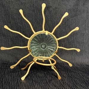May include: A gold-colored metal candle holder with a sunburst design. The center features a clear, textured glass insert. The candle holder has a circular base and multiple curved arms extending outwards, with a small ring for a candle.