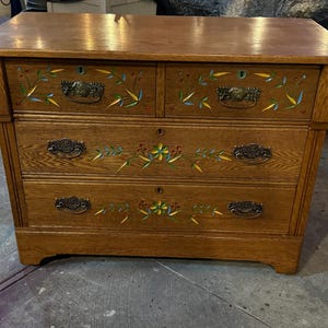 May include: A wooden dresser with a natural brown finish featuring three horizontal drawers. The front of the drawers is decorated with hand-painted floral motifs in yellow, blue, and green. Each drawer is equipped with dark metal pull handles.