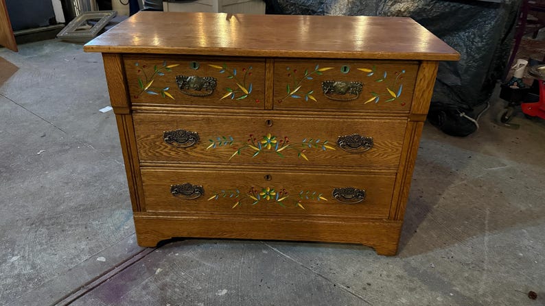 May include: A wooden dresser with a natural brown finish featuring three horizontal drawers. The front of the drawers is decorated with hand-painted floral motifs in yellow, blue, and green. Each drawer is equipped with dark metal pull handles.