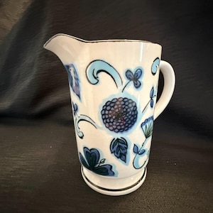 May include: A white ceramic pitcher featuring a hand-painted floral pattern in various shades of blue, including light blue swirls and dark blue blossoms. The pitcher has a curved handle, a flared pouring spout, and a silver-toned rim and base.