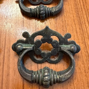 May include: A set of dark metal drawer pulls featuring a decorative backplate with a cutout floral pattern and a curved, ribbed drop handle. The metal has a weathered, aged finish with visible patina and subtle green oxidation on the surface.
