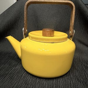 Puede incluir: Una tetera de color amarillo brillante con asa y tapa de madera. La tetera tiene un pico curvo y un cuerpo redondeado. El asa es de madera clara y está sujeta con soportes metálicos. La tapa tiene un pequeño pomo de madera.