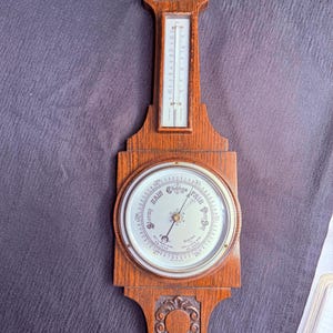 May include: A vintage wooden barometer and thermometer combination. The barometer has a circular face with a needle indicating weather conditions. The thermometer is a vertical glass tube. The wooden frame has decorative carvings.