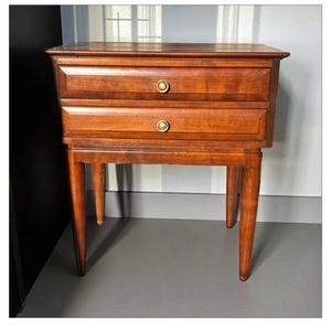 May include: A two-drawer wooden bedside table in a warm, reddish-brown finish. The drawers have round, brass-coloured knobs. The table has four tapered legs.