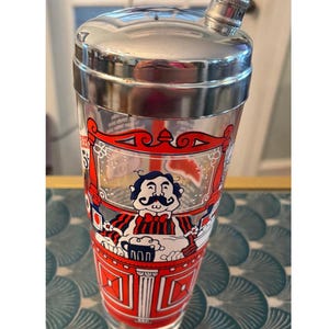 Puede incluir: Coctelera vintage con tapa plateada y cuerpo de vidrio transparente. El vidrio presenta un diseño rojo y blanco con una imagen de dibujos animados de un hombre con bigote. La coctelera está diseñada para mezclar bebidas.