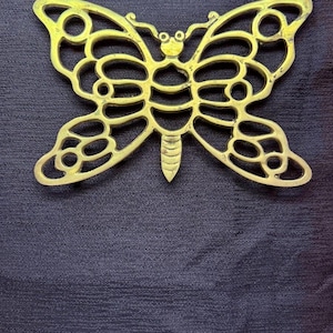 Puede incluir: Un salvamanteles de metal dorado con forma de mariposa. El salvamanteles tiene un diseño calado con recortes redondeados y un cuerpo detallado. Se muestra sobre una superficie texturizada de color azul oscuro.