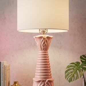 May include: A pink ceramic table lamp with a cream-colored lampshade. The lamp has a decorative base with a fluted design and a stacked, ribbed column. The lamp is approximately 28 inches tall.