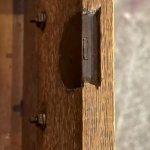 May include: A light brown oak wood panel featuring a recessed dark metal latch mechanism embedded into the side edge. Two small metal bolts are visible on the flat surface of the wood, which displays a prominent vertical grain pattern.