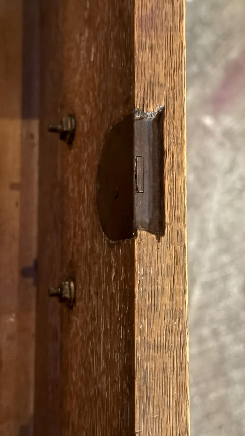 May include: A light brown oak wood panel featuring a recessed dark metal latch mechanism embedded into the side edge. Two small metal bolts are visible on the flat surface of the wood, which displays a prominent vertical grain pattern.