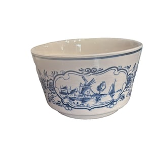 Puede incluir: Un cuenco de cerámica blanco con un diseño de molino de viento holandés azul y blanco. El cuenco tiene una forma curva y un borde azul. El diseño presenta un molino de viento, casas y un barco. El cuenco probablemente se utiliza para servir o exhibir.