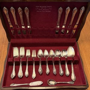 Puede incluir: Un juego de cubiertos plateados en un estuche de madera oscura con interior de terciopelo rojo intenso. El conjunto incluye varios tenedores, cucharas y cuchillos, con el texto 1847 Rogers Bros. 1847 Anniversary grabado en la tapa del estuche.