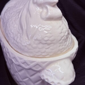 May include: A white ceramic hen-shaped serving dish with a textured surface. The lid is shaped like a hen, and the base has a basket weave design. The dish is likely used for serving food.