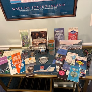 May include: A collection of vintage memorabilia, including brochures, maps, and a commemorative cup, related to the New York World's Fair. The items are displayed on a glass-topped table, with a framed poster in the background. The poster reads "MADE ON STATEN ISLAND".