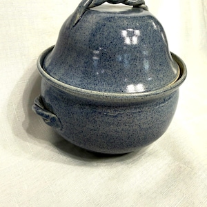 May include: A blue ceramic lidded bowl with a speckled finish. The lid has a twisted handle, and the bowl has small handles on the sides. The bowl is sitting on a white surface.