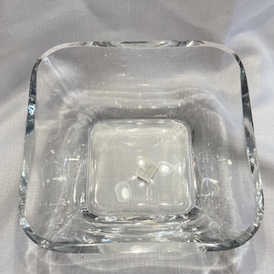 May include: A clear, square glass bowl with thick, rounded edges and a smooth, transparent surface. The bowl features a deep, open interior and a small, square white sticker attached to the bottom center of the basin, visible through the glass.