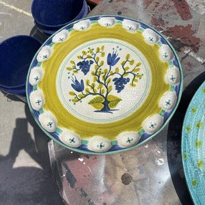 Könnte beinhalten: Ein dekorativer Keramikteller mit einem gelb-weißen, gewellten Rand. Die Mitte zeigt ein Baumdesign mit blauen Blüten und grünen Blättern. Der Teller hat einen Durchmesser von ca. 25 cm.