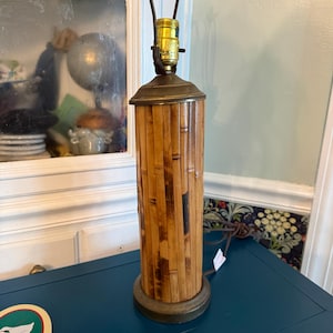 Puede incluir: Una lámpara de mesa vintage con una pantalla cilíndrica de bambú y detalles en latón. La lámpara tiene un cuerpo de bambú marrón, una base de latón y una parte superior de latón. La lámpara está sobre una superficie azul.