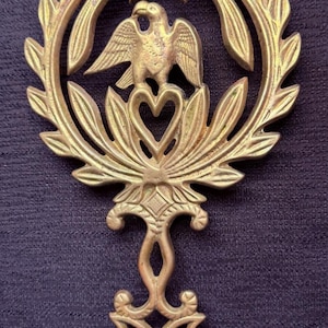 Könnte beinhalten: Ein goldfarbenes Metall-Dekoremblem mit einem Adler mit ausgebreiteten Flügeln, umgeben von einem Lorbeerkranz. Das Design umfasst ein herzförmiges Element und eine dekorative Basis, vor einem dunkelblauen, strukturierten Hintergrund.