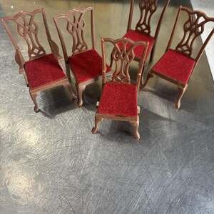 May include: Six miniature brown chairs with red velvet seats. The chairs have decorative backs and are arranged on a metallic surface. These chairs are likely for a doll's house or miniature display.