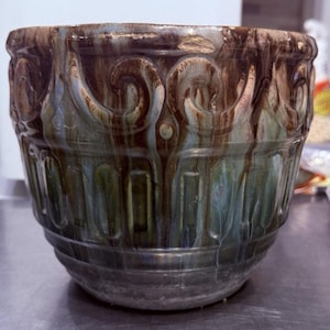 Può includere: Una ciotola in ceramica con finitura lucida, caratterizzata da una miscela di tonalità marroni, verdi e blu. La ciotola ha un design ornato e strutturato con motivi ripetuti e una forma arrotondata. L'esterno ha un effetto smaltato a goccia.