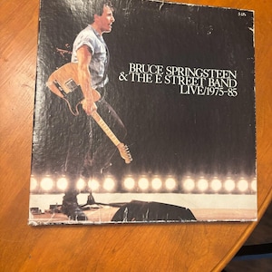 Puede incluir: Una portada de álbum de vinilo vintage con Bruce Springsteen & The E Street Band. La portada del álbum es negra con una foto en vivo de Springsteen tocando la guitarra. El texto en la portada dice "BRUCE SPRINGSTEEN & THE E STREET BAND LIVE/1975-85."