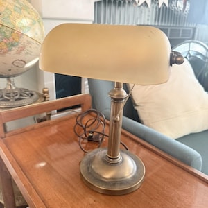 May include: A banker's lamp with a brass-toned metal base and a cylindrical, frosted glass shade. The lamp features a vertical, ribbed central column, a round base, and a pull chain switch. A brown electrical cord is coiled at the base.