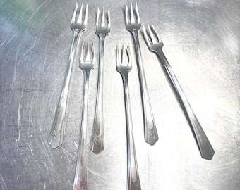 Op de afbeelding: Zes zilveren vorken met elk drie tanden, gerangschikt op een metalen oppervlak. De vorken hebben lange, slanke handvatten met een decoratief ontwerp aan het uiteinde. Keukengerei.
