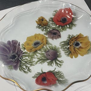Puede incluir: Una fuente de vidrio transparente con borde dorado y borde ondulado. La fuente está decorada con diseños florales coloridos, incluyendo anémonas rojas, amarillas y moradas con follaje verde.