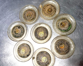 May include: A collection of eight clear, round coasters with a steampunk design. Each coaster features a central, detailed gear mechanism in gold and bronze tones, encased in a clear, slightly textured border. The coasters are arranged on a metallic surface.
