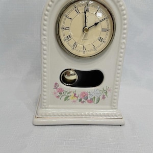 May include: A white ceramic quartz clock with a cream-colored face and black Roman numerals. The clock has a decorative floral design and a small, round, mirrored accent. The clock is approximately 6 inches tall.