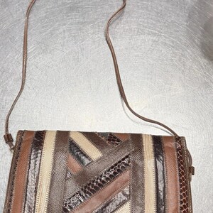 May include: A rectangular shoulder bag with a patchwork design. The bag features a variety of brown, tan, and cream-colored leather pieces stitched together. The strap is a thin, matching brown cord. The bag is set against a metallic surface.