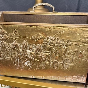 May include: A rectangular, antique brass magazine rack with a handle. The front panel features a detailed relief scene of a horse-drawn carriage, people, and buildings. The interior is lined with a dark brown material.
