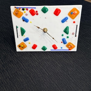 May include: Square white clock with colorful glass accents. The clock face features red, green, blue, and orange glass pieces around the perimeter. The clock hands are gold-colored. A decorative piece for a home or office.