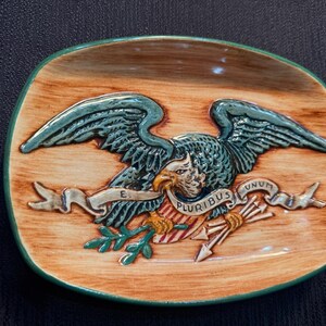 Peut inclure: Un plat décoratif en céramique avec un fond marron imitation bois et un bord vert. Le plat présente un aigle détaillé de couleur sarcelle aux ailes déployées, tenant une bannière avec l'inscription "E. PLURIBUS UNUM".