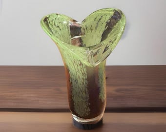 May include: A decorative glass vase with a flared, petal-like top. The vase features a green and brown speckled design. The vase is approximately 20 cm tall and has a dark base. The vase is sitting on a wooden surface.